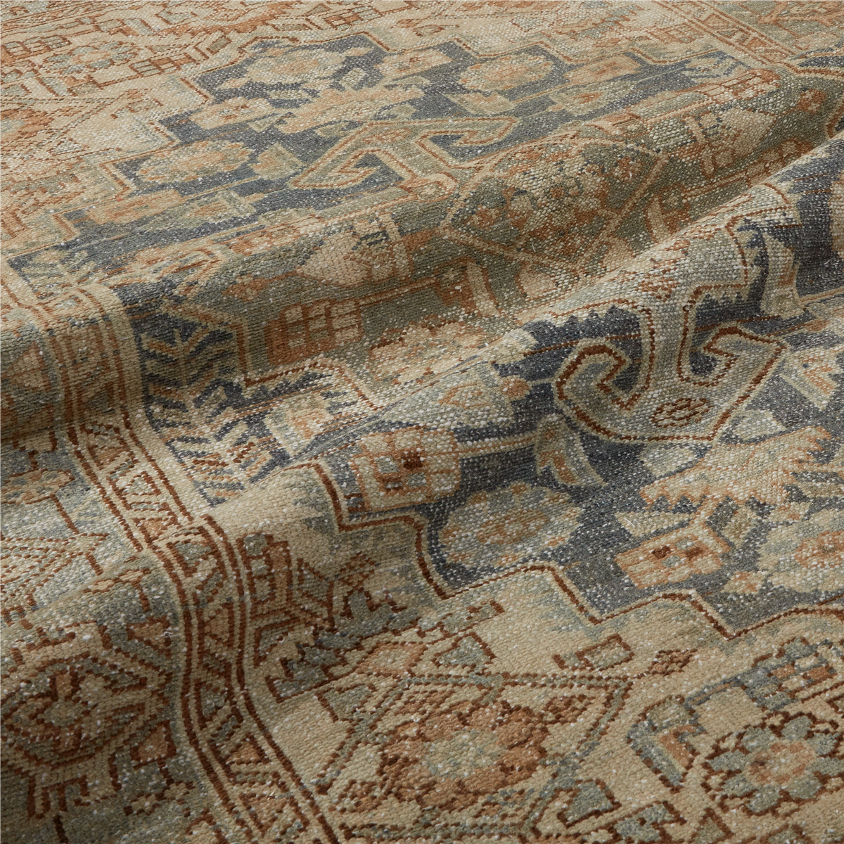 A close-up of a traditional patterned rug with a distressed, vintage aesthetic. The rug features a complex geometric and floral motif in muted tones of dusty blue, warm beige, terracotta, and brown.