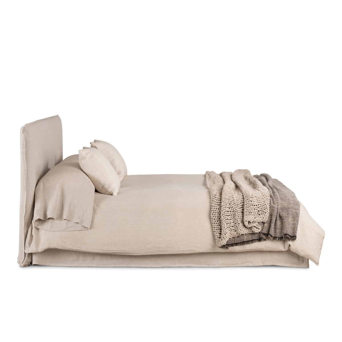 Muse Queen Bed in side profile in natural linen, with a tall slipcovered headboard and a skirted base, styled with linen bedding and a chunky knit throw.