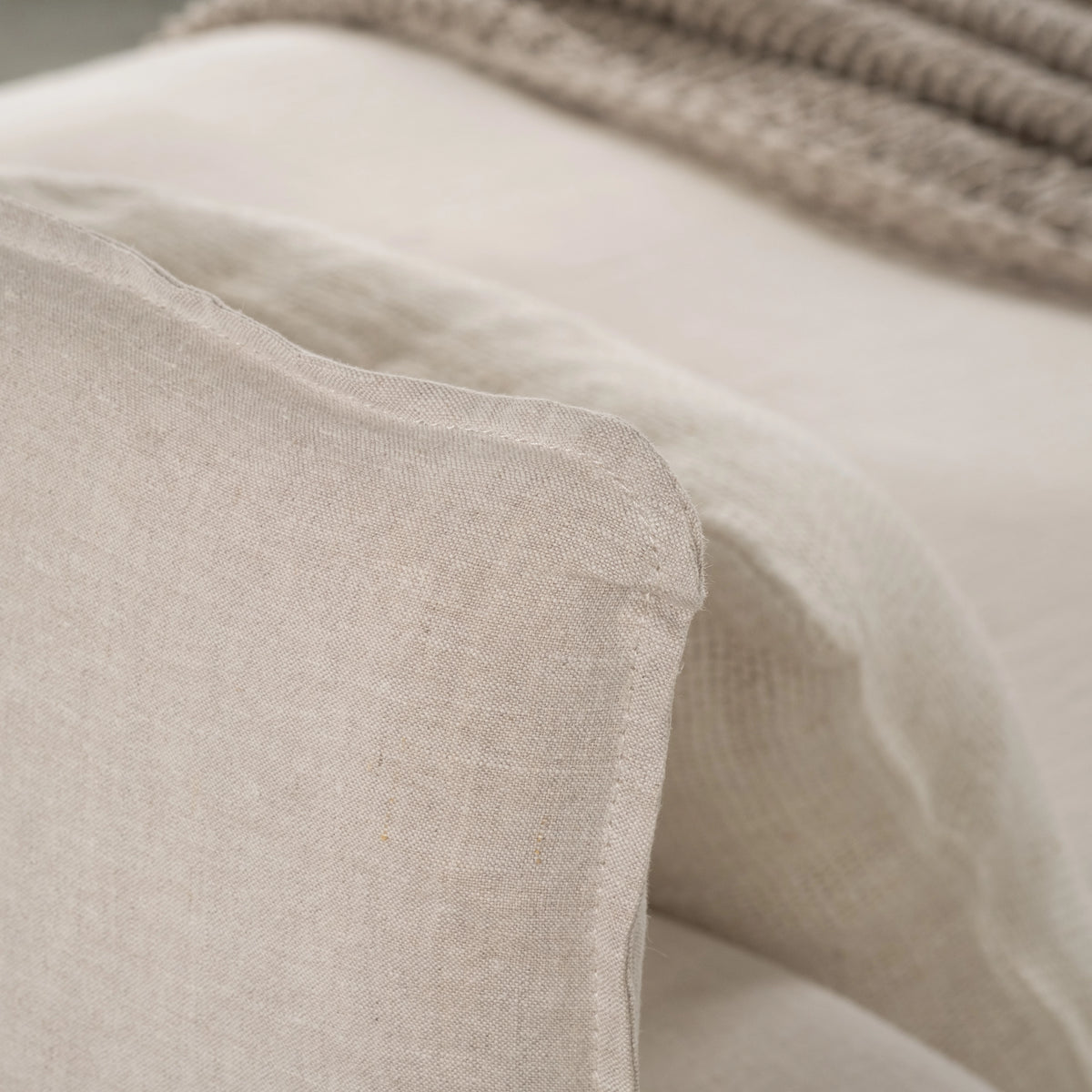 Detail of the Muse Queen Bed's linen headboard edge and stitched seam, with linen bedding and a knit throw softly out of focus behind.
