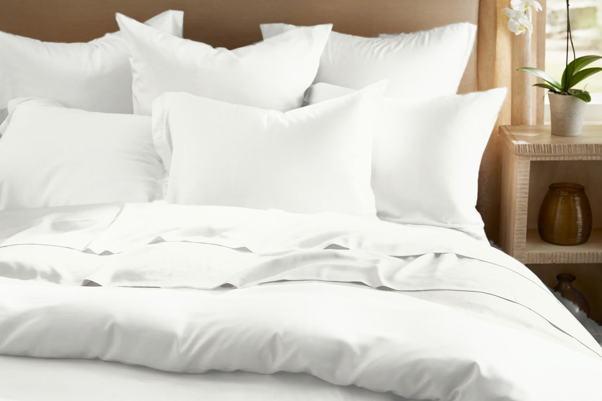 Legna Classic Bedding Set in Cloud, a bright white with a subtle sheen, shown from the front with layered euro and lumbar pillows, a crisp flat sheet fold, and a full duvet cover beside a wood nightstand.
